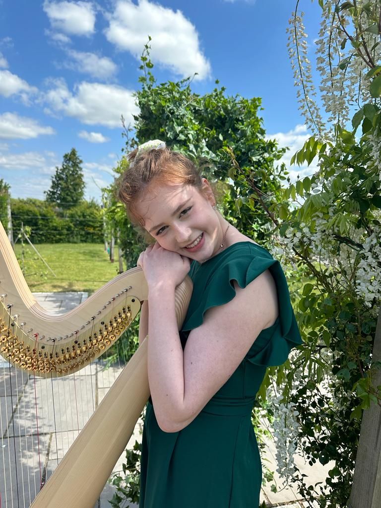Sebrina in a green dress leaning on a harp outdoors in a sunny garden with flowers.