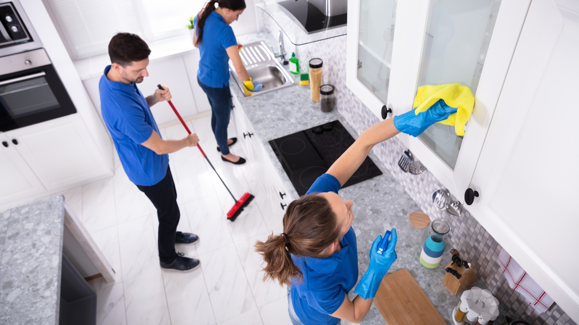 Three people in blue shirts cleaning a kitchen: sweeping, wiping countertops and cabinets.