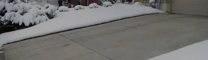 Snow-covered driveway next to bushes and a garage. Winter scene with concrete pavement and a snow bank.
