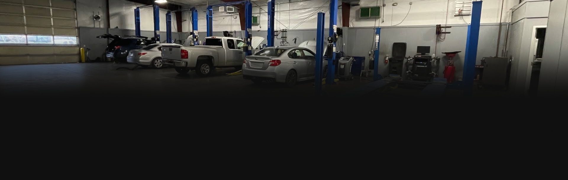 Inside a vehicle repair shop, cars and a pickup truck are on lifts, garage door open.