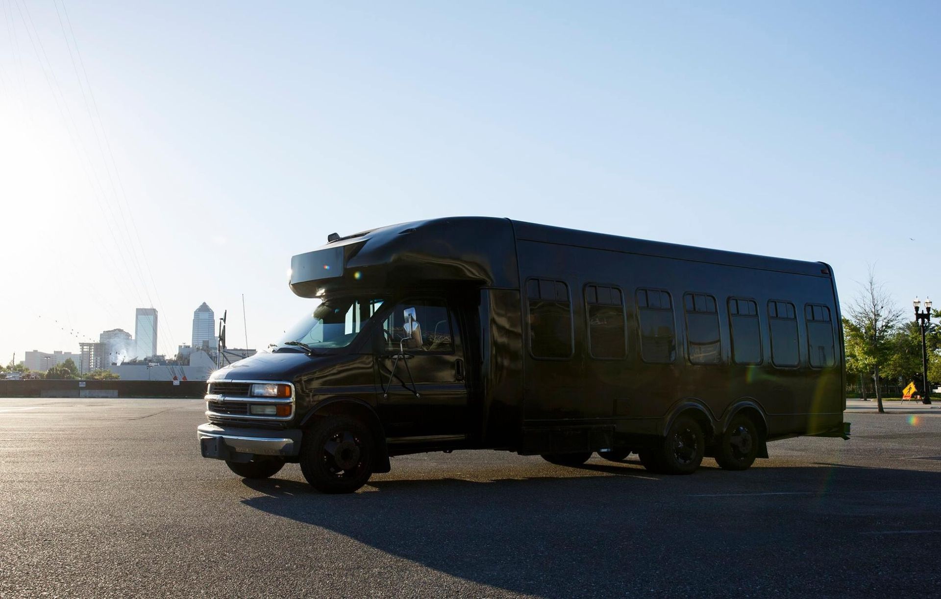 Dark bus parked on open pavement with city skyline in the background at sunset