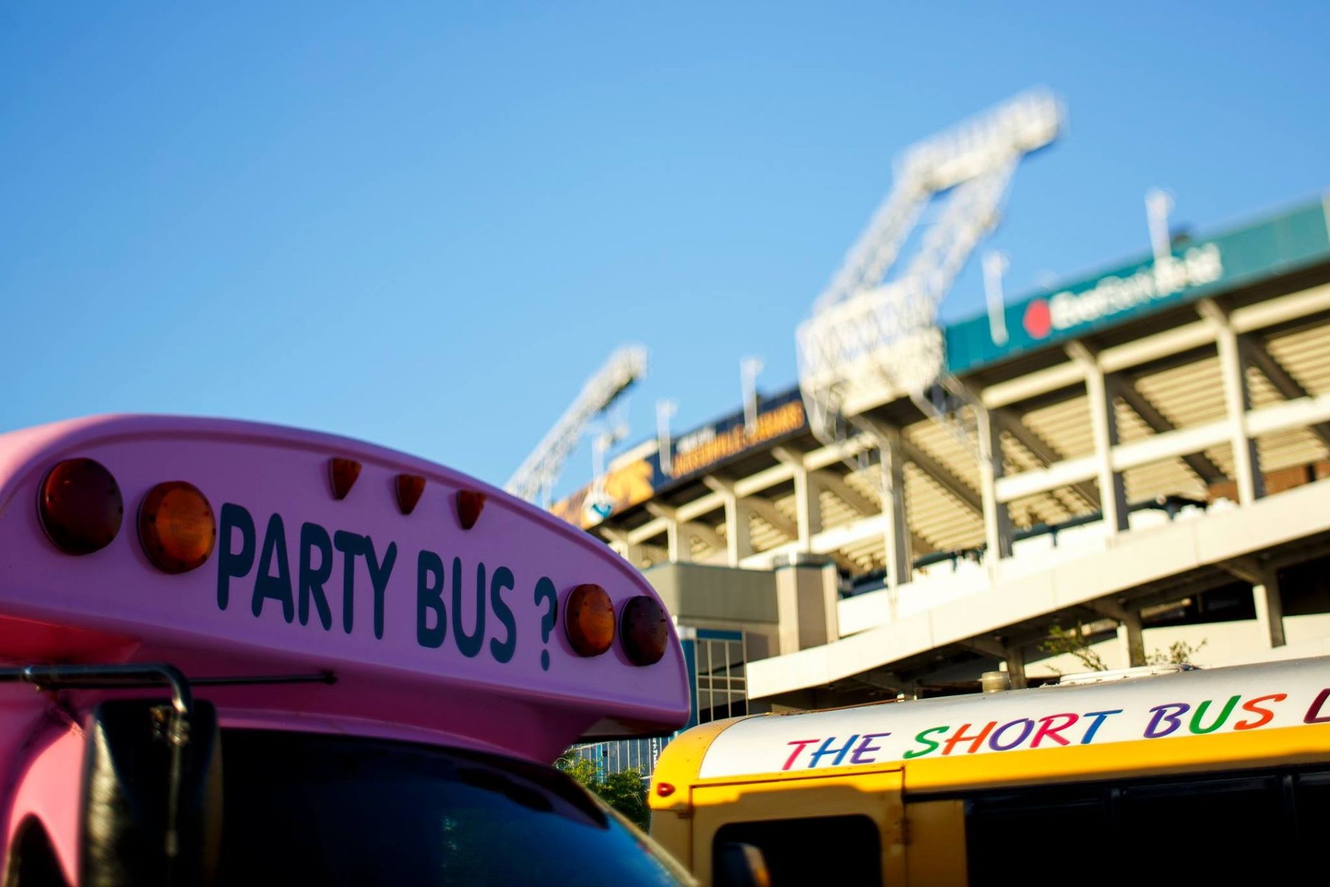 Two colorful buses at a stadium, including a pink Party Bus and a yellow Short Bus under blue sky