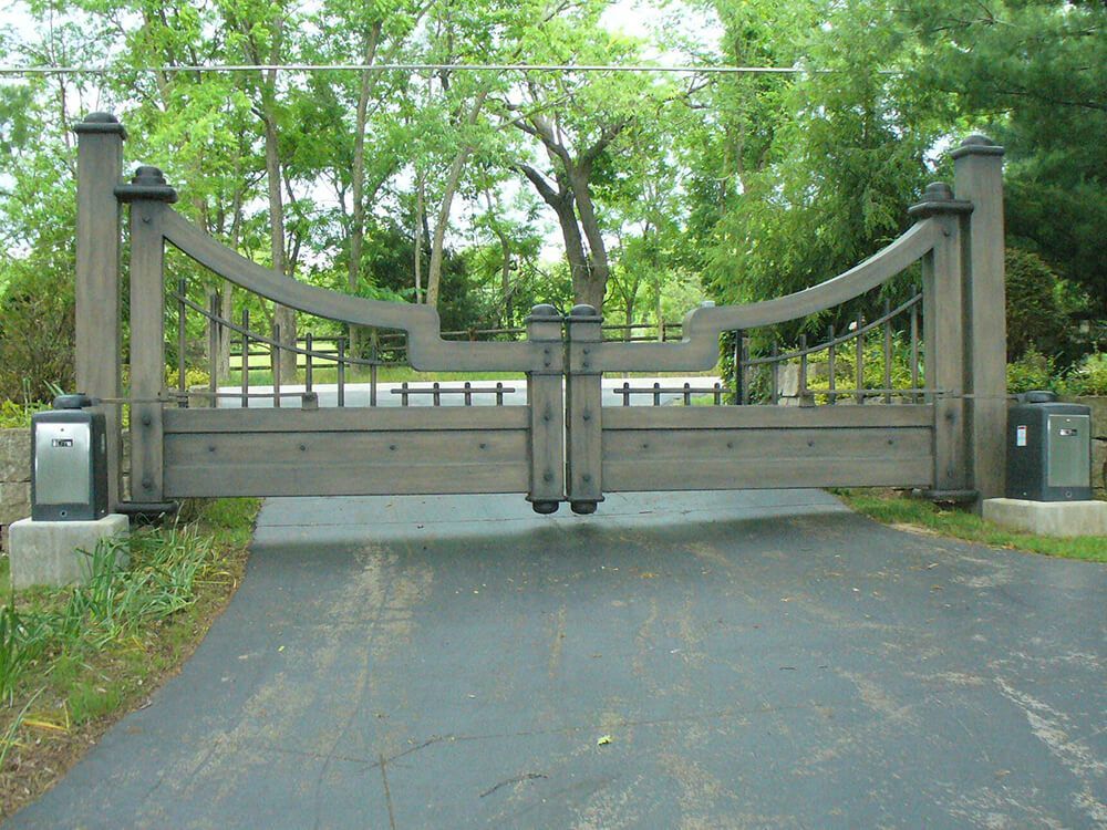 A wooden gate is open to a driveway with trees in the background