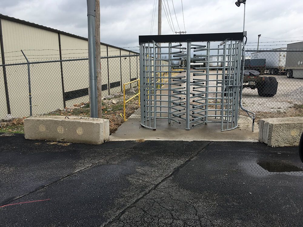 A turnstile in a parking lot with a chain link fence in the background