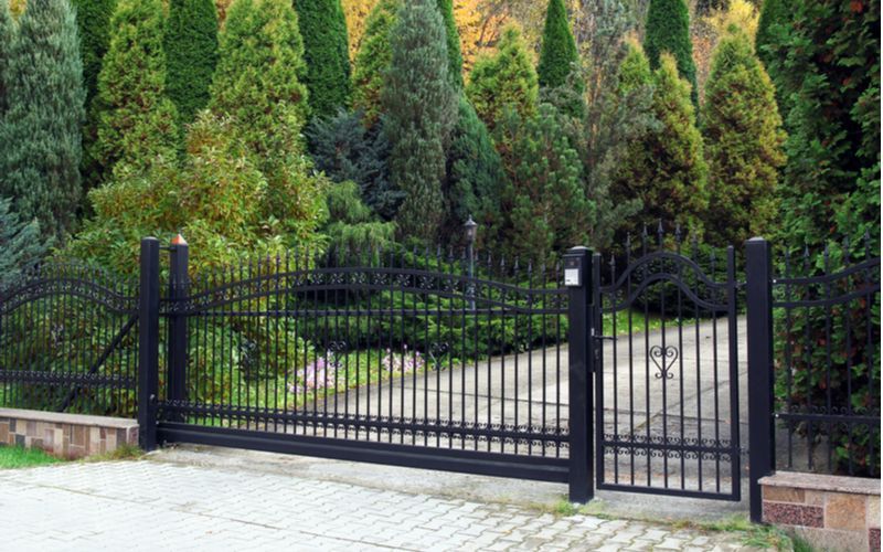 A black fence is surrounding a driveway with trees in the background.