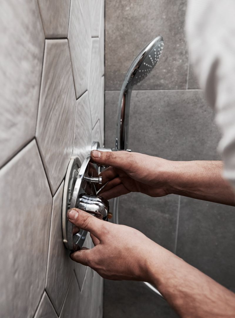 Person's hands working on a chrome shower fixture against a gray tiled wall.