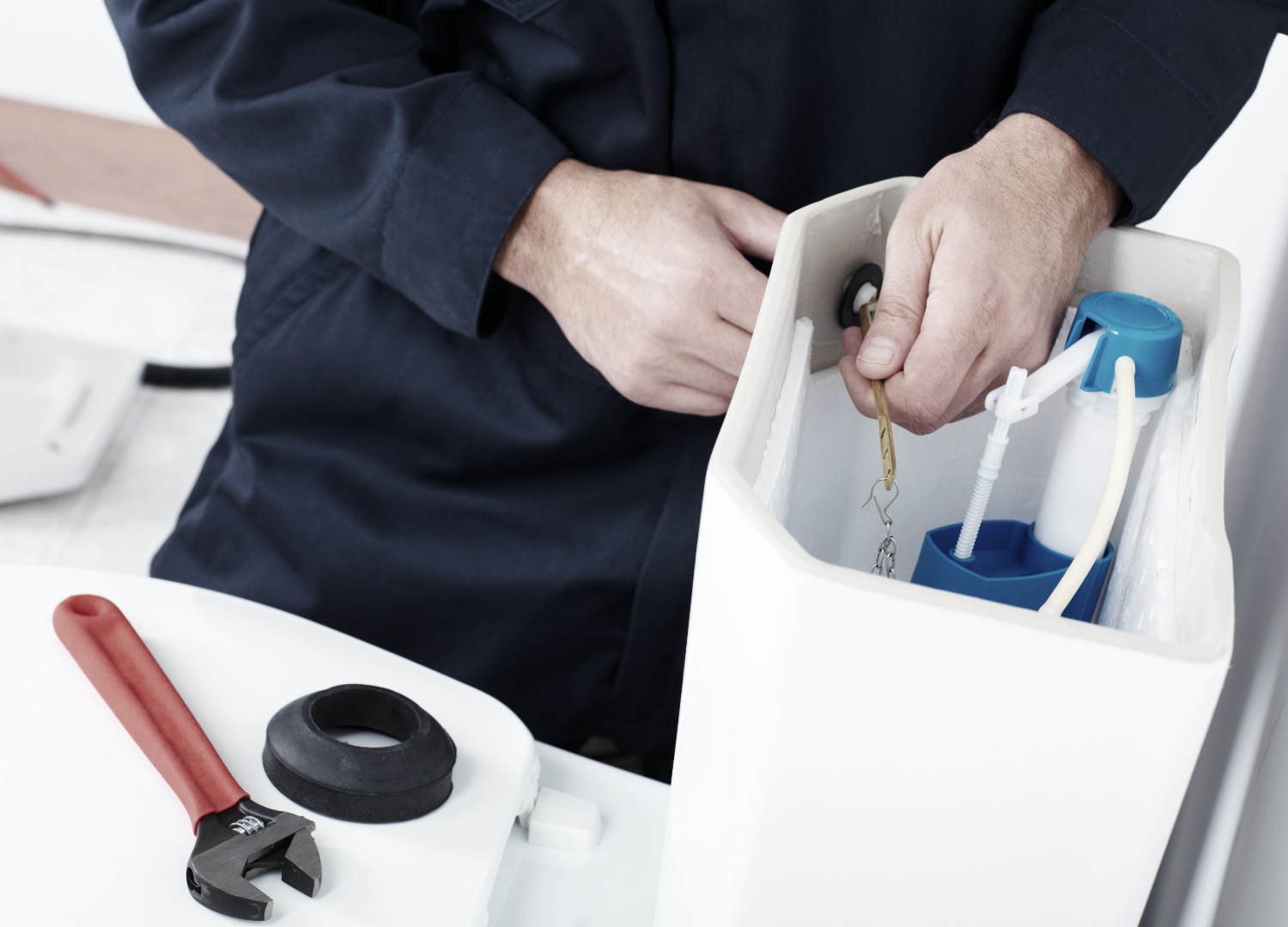 Plumber in navy jumpsuit repairing a toilet. He's holding internal parts, a wrench and black seal rest on the toilet lid.