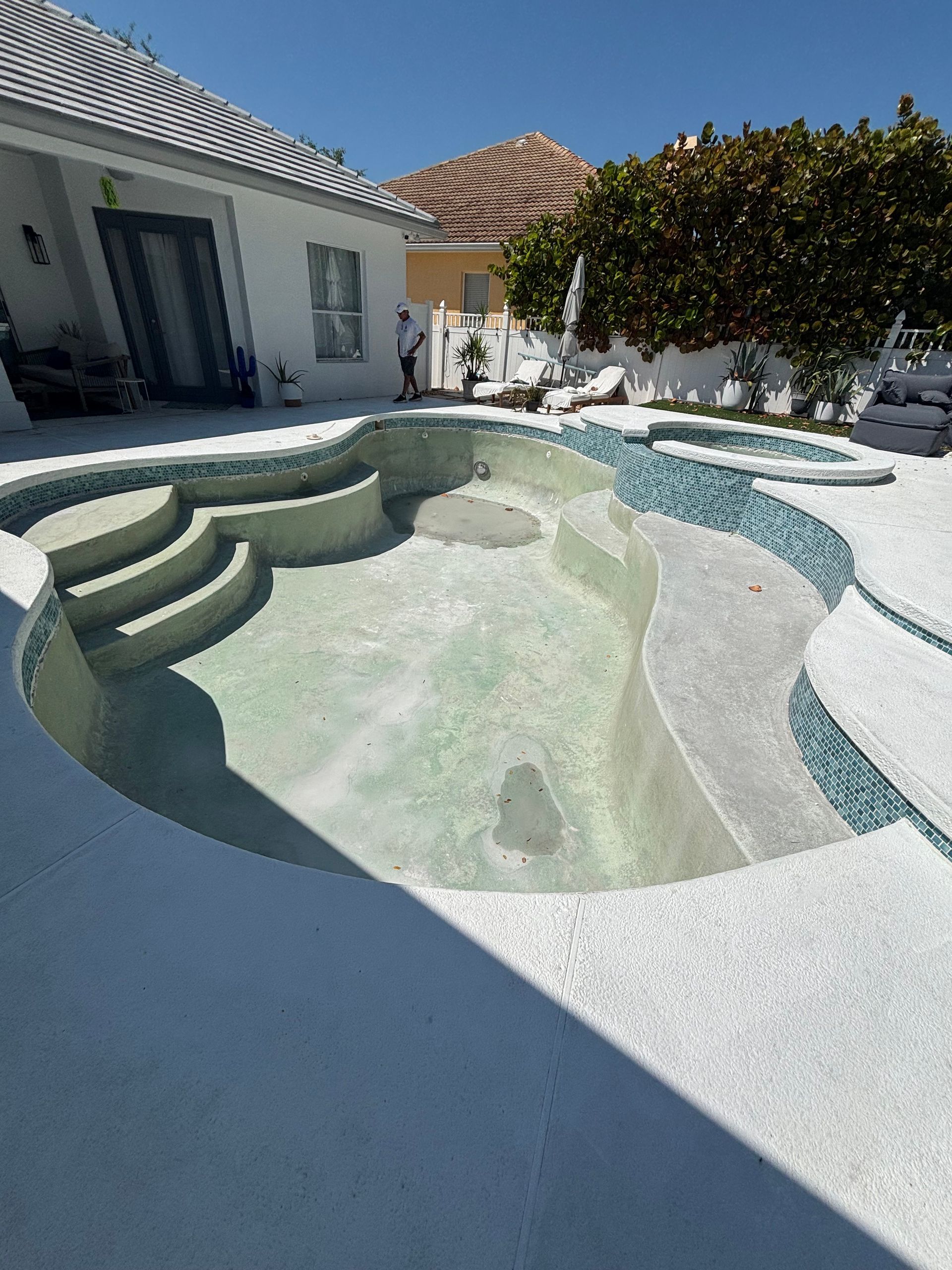 Empty white backyard pool with built-in steps and a nearby house and trees under bright sun