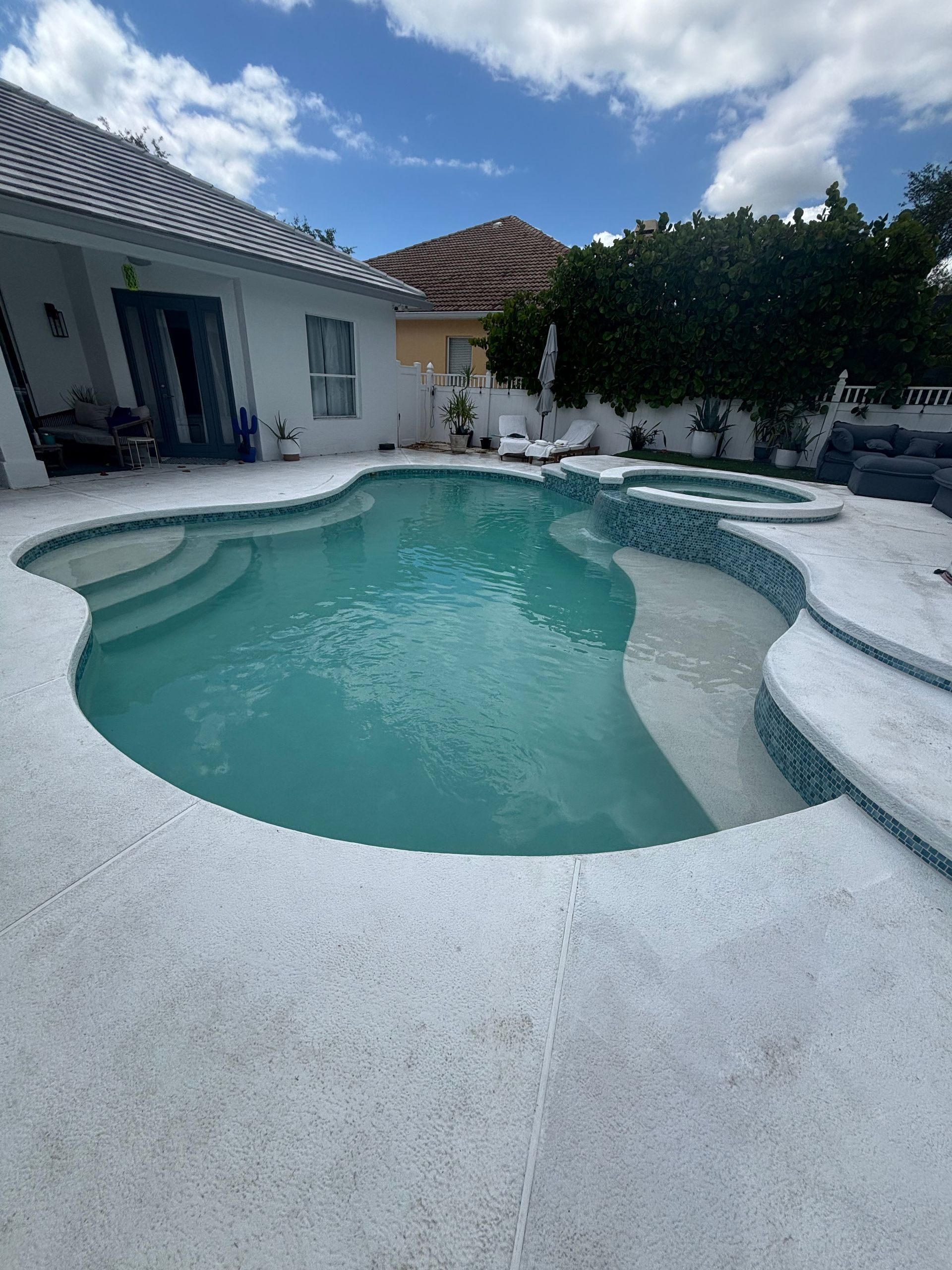 Backyard pool with white stone decking, patio house, and cloudy sky