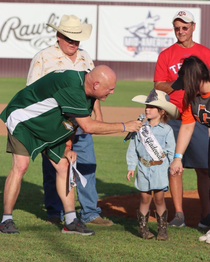 Adult hands child a sash at a baseball field ceremony, with others watching nearby.