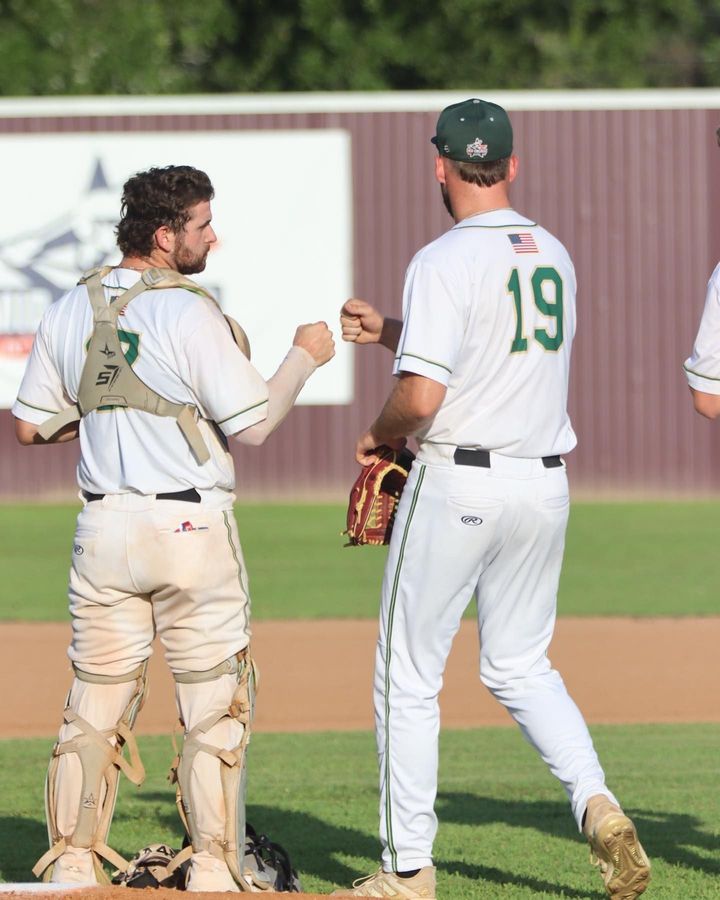 Two baseball players in white uniforms fist bump on the field near the outfield fence.