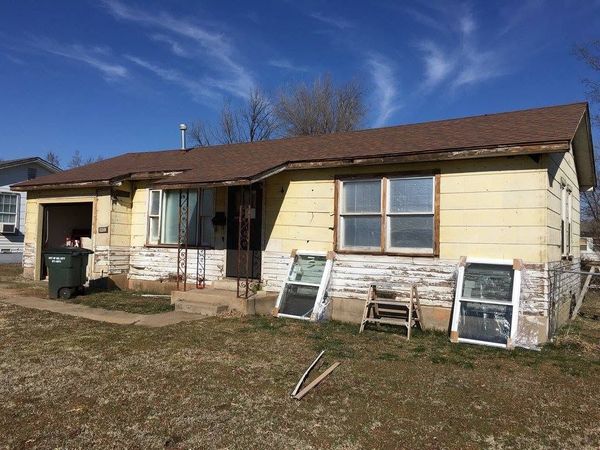 Small weathered yellow house with a brown roof and cluttered front yard under a blue sky