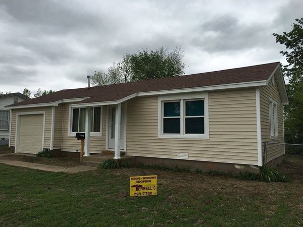 Single-story beige house with brown roof under cloudy sky, with a yellow yard sign in front.