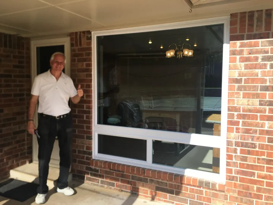 Man standing on a brick porch beside a large window, wearing a white shirt and dark pants.