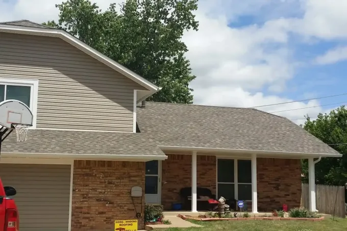 Suburban house with brick and beige siding, a front porch, basketball hoop, and a red vehicle at left.