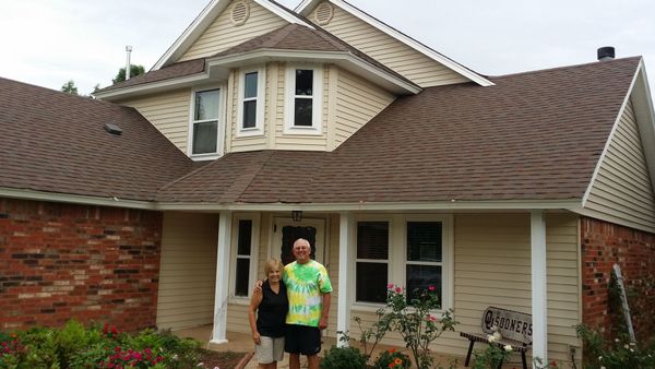 Two people standing in front of a brick house with a brown roof and a small porch.