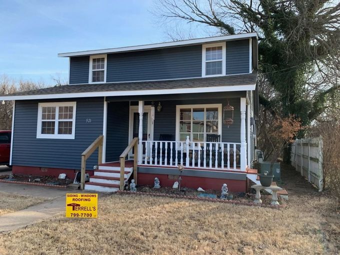 Two-story blue house with white trim and a front porch, viewed from a dirt yard.
