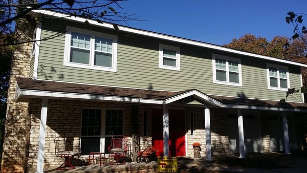 Two-story green house with white trim, front porch, and red chairs in fall sunlight