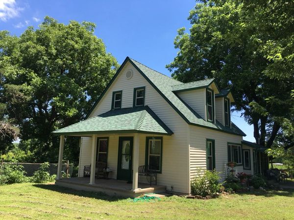 Small cream-colored house with green roof and porch surrounded by trees under a blue sky