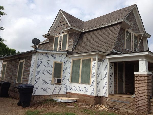 Two-story house under renovation with white wrap, brick porch, and black trash bins outside