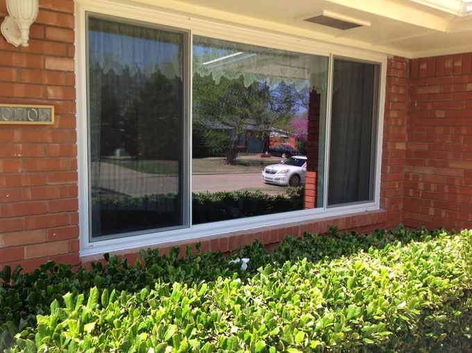 Brick building window reflecting trees and a parked white car, with green shrubs below