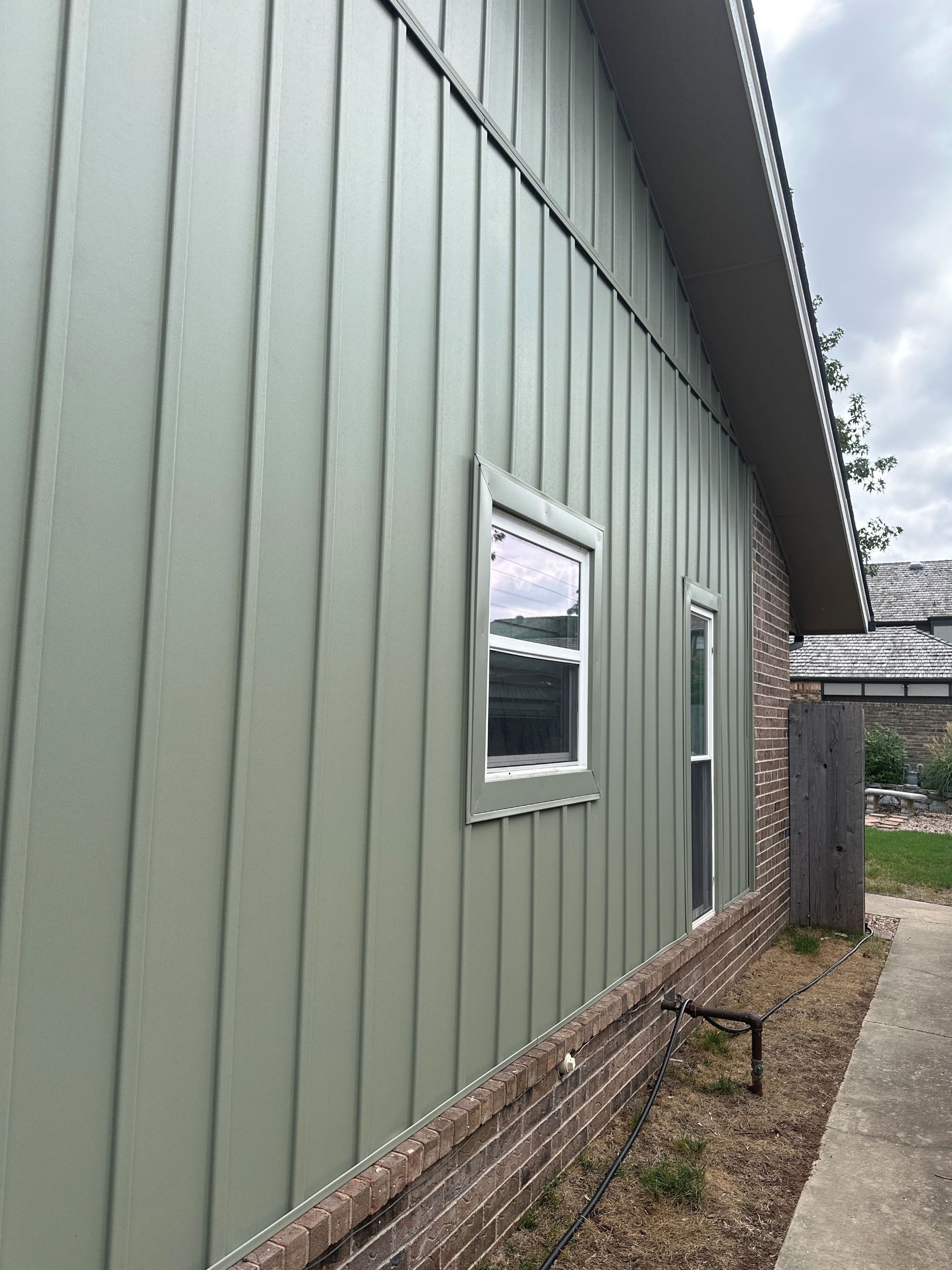 Green metal-sided building with a small window beside a gravel path and brick foundation