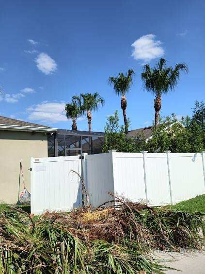 White fence with debris in front of a house, palm trees in the background under a blue sky.