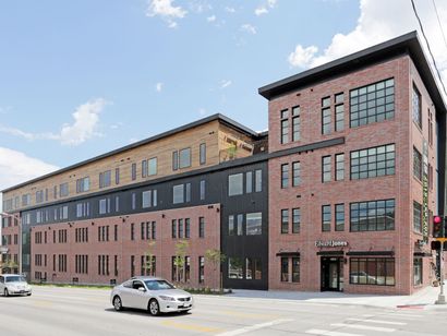 A white car is driving down the street in front of a large brick building.