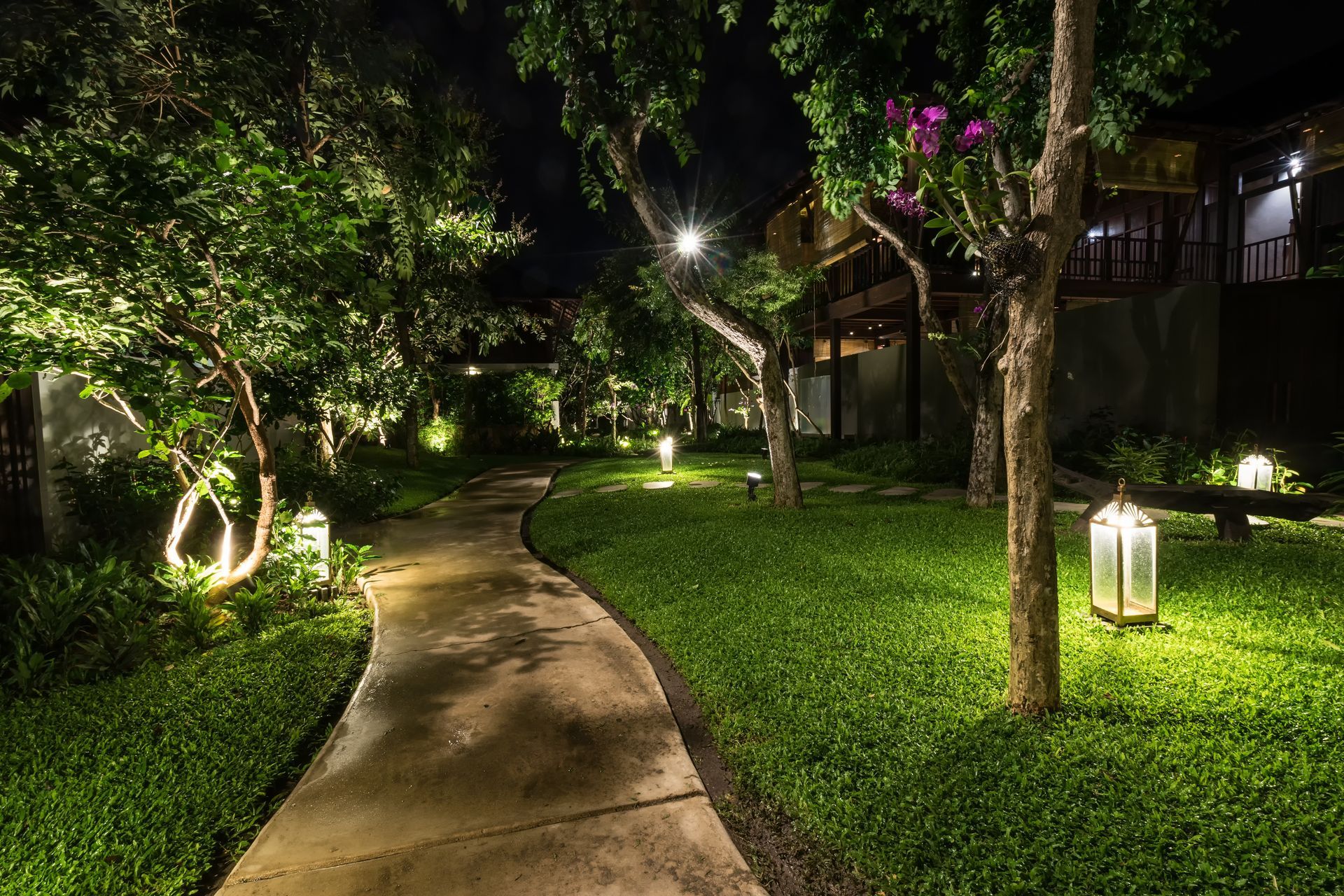 Curved garden path lit by warm lanterns among lush green trees and grass at night