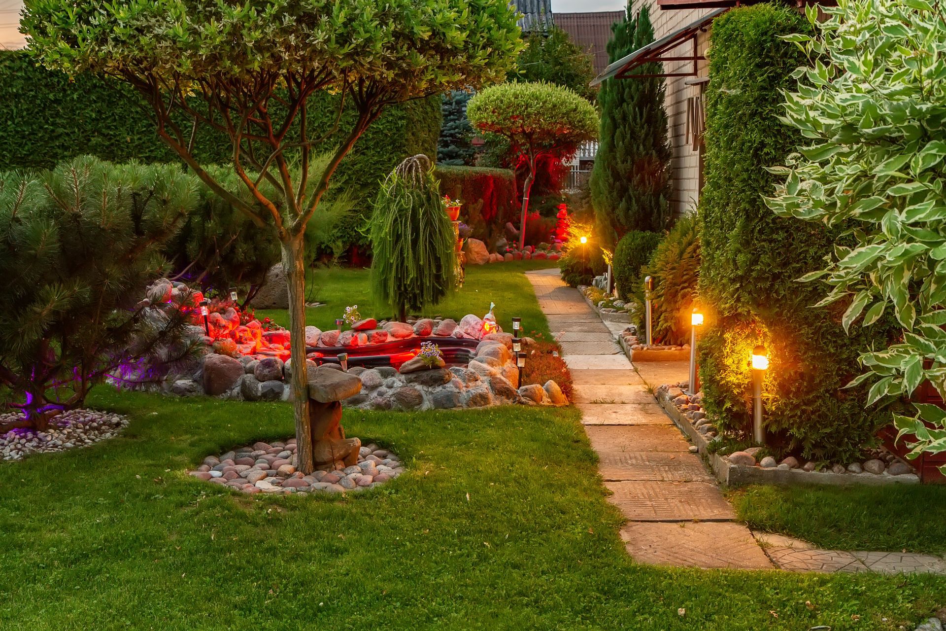 A lit garden path with stone steps, green lawn, and flowering shrubs at dusk.