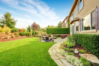Backyard patio beside a house with green lawn, stone path, and garden beds under a blue sky