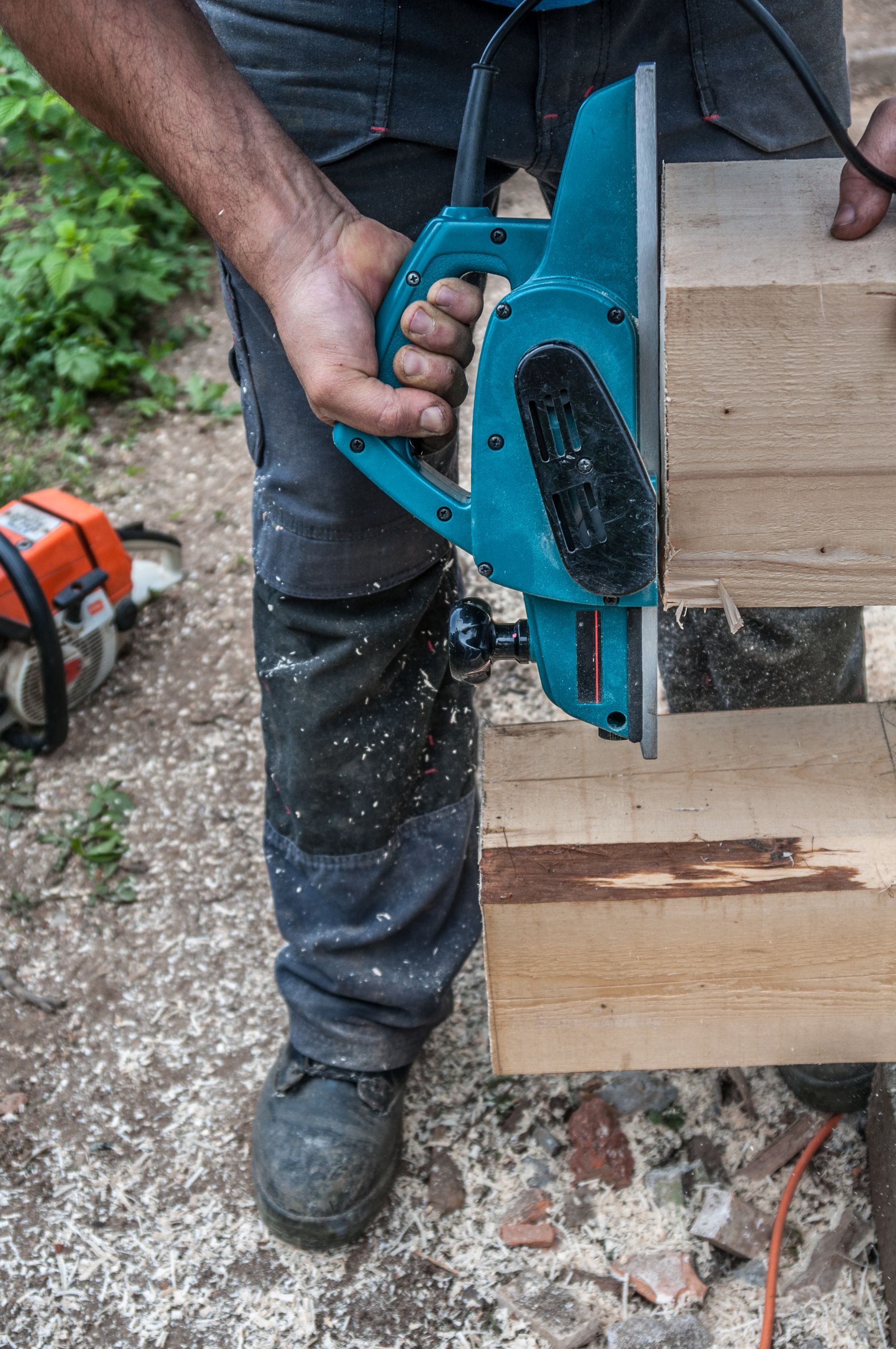 Man using an electric planer on a wooden beam outdoors. Cyan tool, wood shavings, and a chainsaw visible.