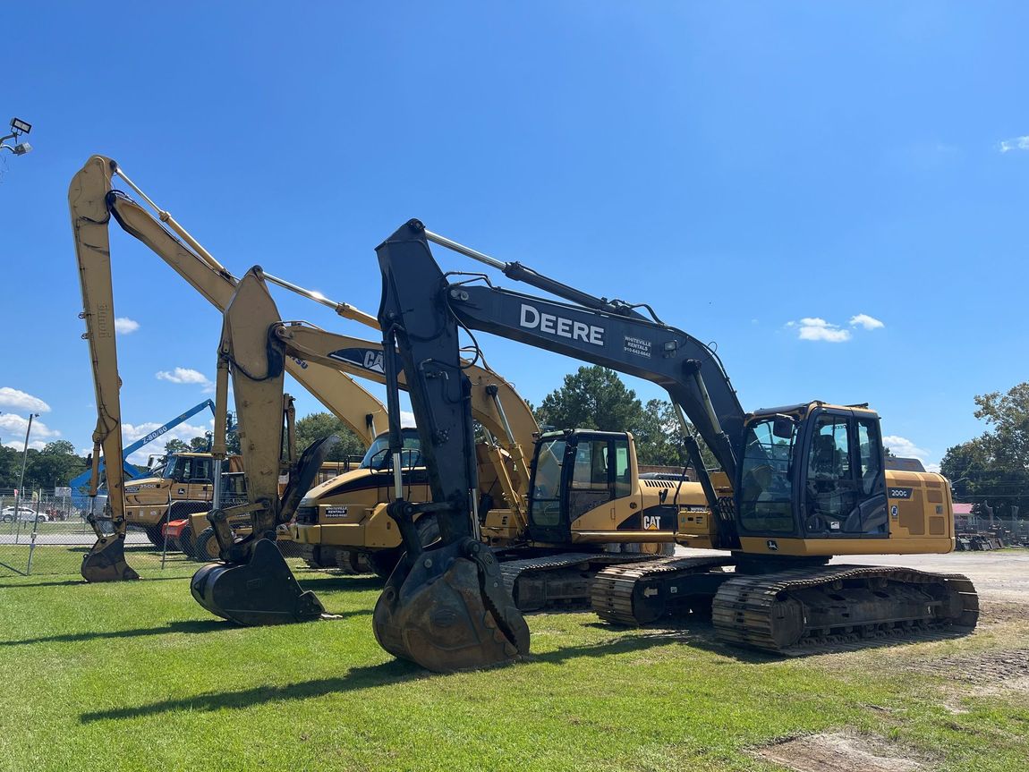 Construction equipment, including excavators, parked on green grass under a blue sky.