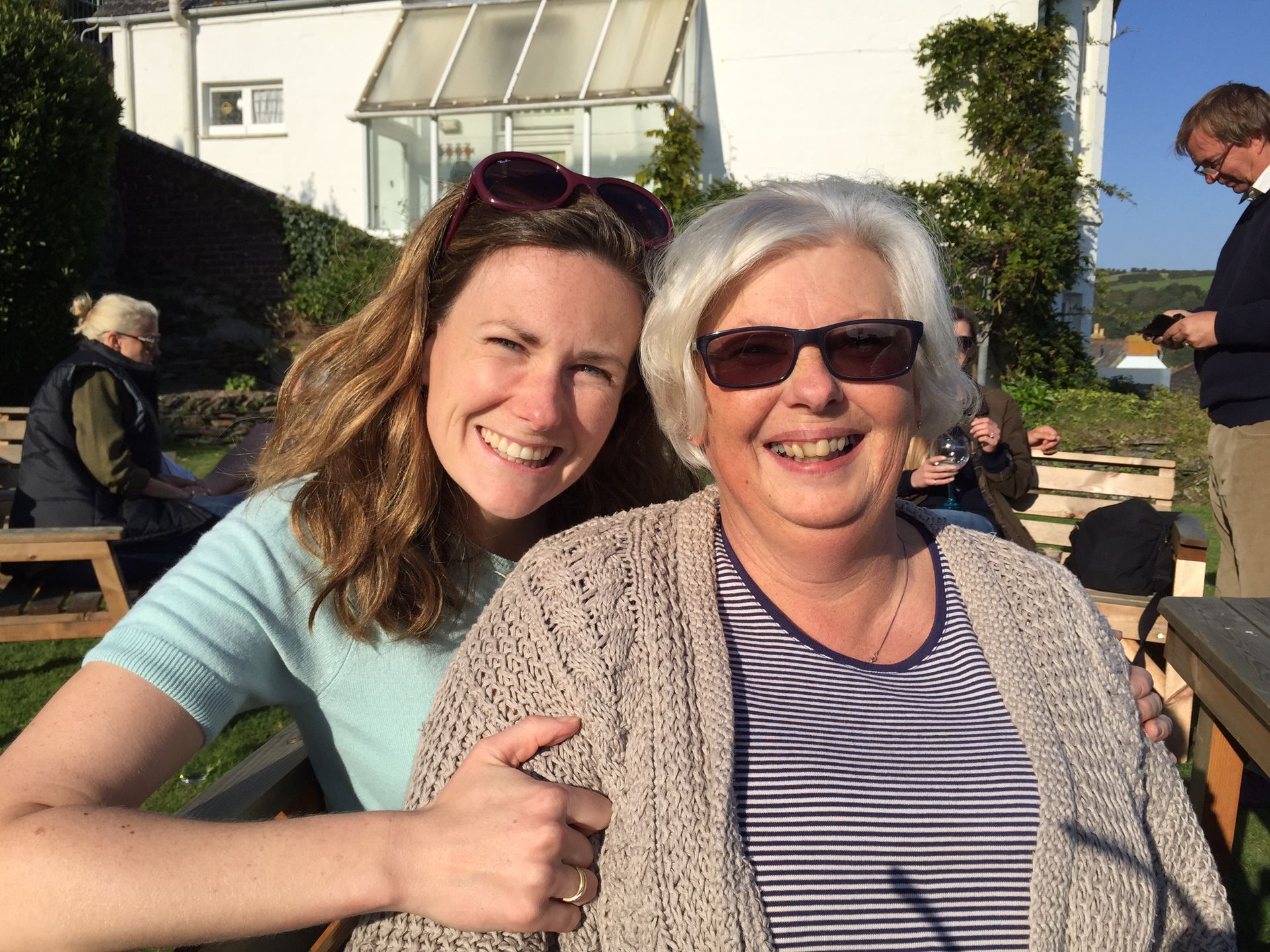 Woman with sunglasses hugs another woman outdoors, both smiling.