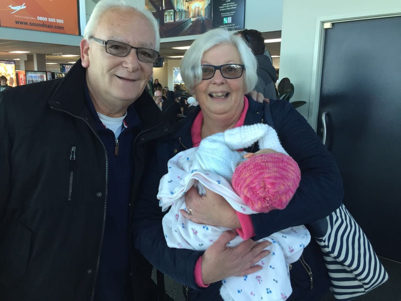 Dr Sam's Parents. Couple holding a baby wrapped in a blanket, smiling at the airport.