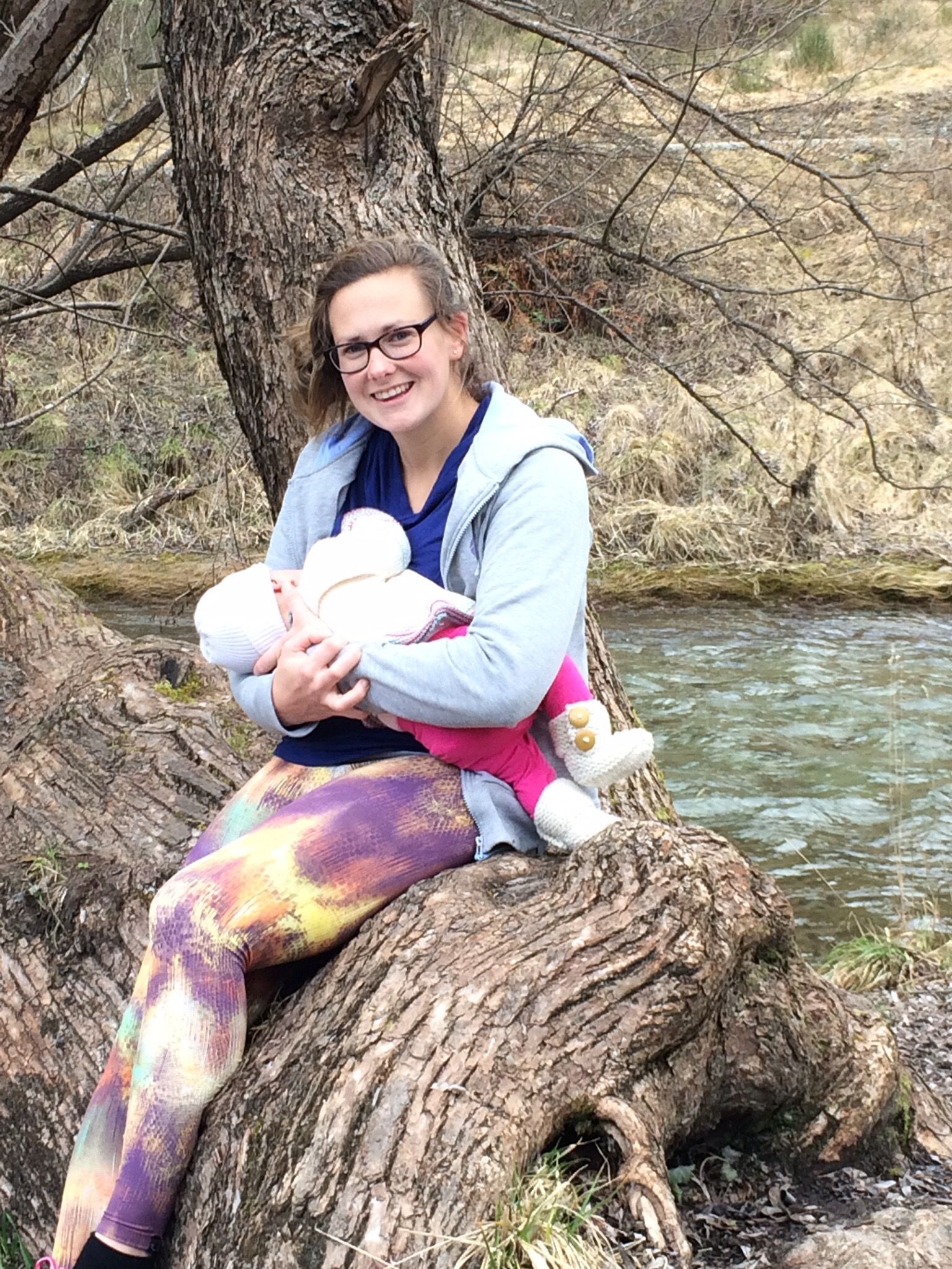Woman in glasses holds baby near a stream, sitting on a tree trunk.