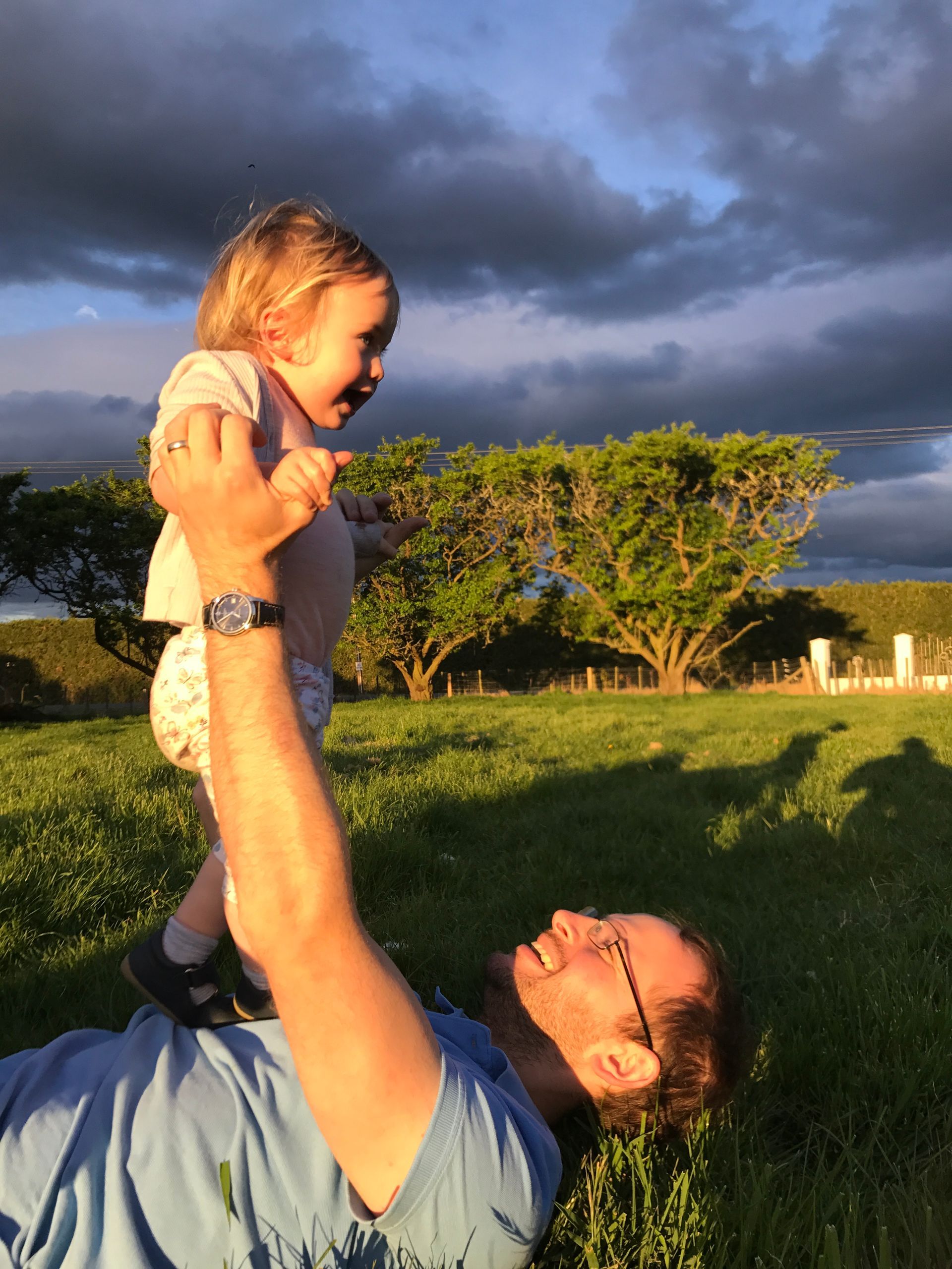 Man lying on grass, holding a small child up in the air. Child is laughing, sky and trees in background.