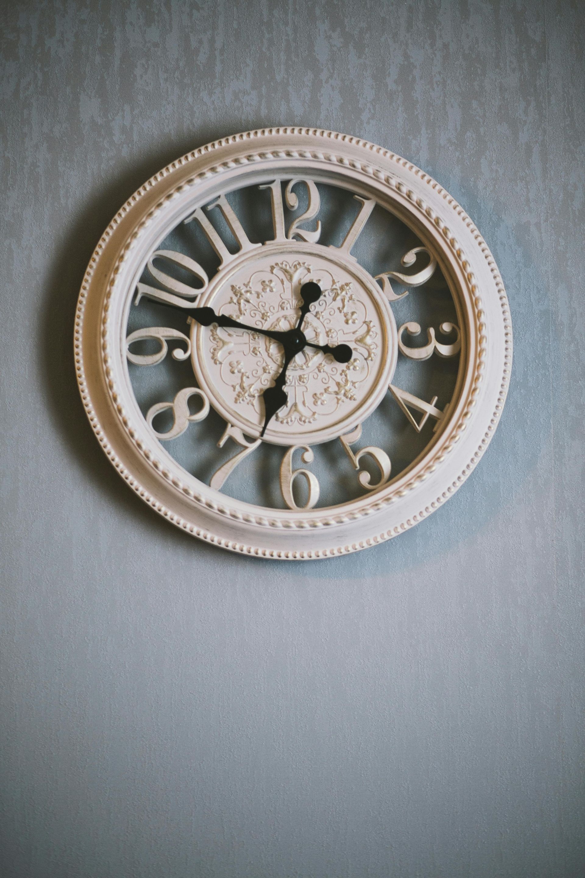 White, ornate wall clock with black hands, mounted on a blue textured wall.