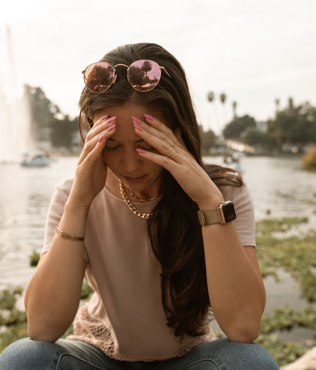 a woman wearing sunglasses is sitting in front of a fountain holding her head .