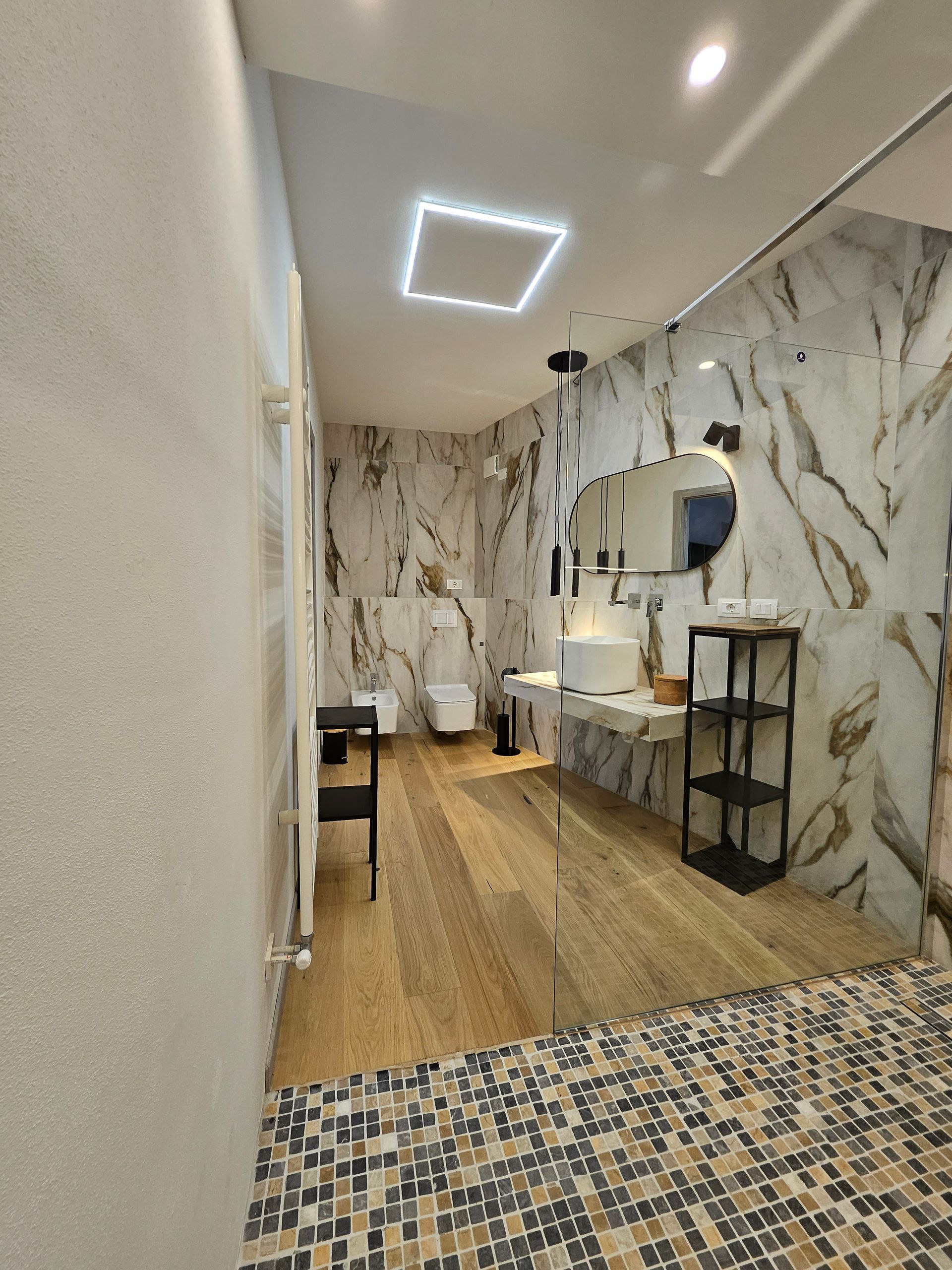 Stretto corridoio del salone rivestito in marmo con pavimento in legno, specchio da trucco e lampadario geometrico a soffitto.