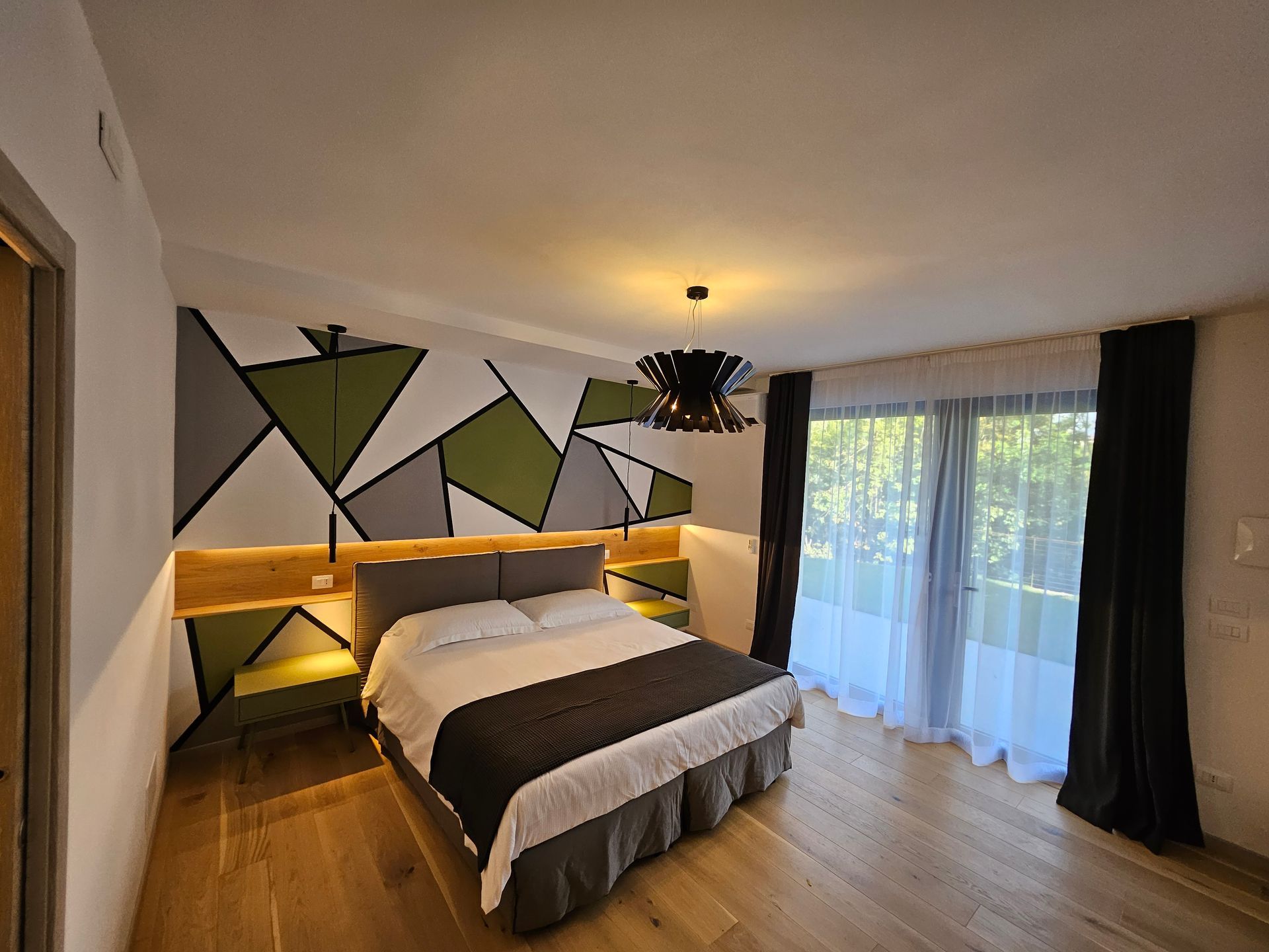 Camera da letto moderna con letto matrimoniale, decorazioni murali geometriche, illuminazione calda e porta-finestra con tende trasparenti.