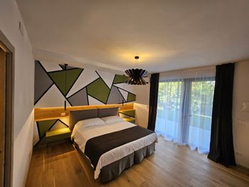 Camera da letto moderna con decorazioni murali geometriche, letto matrimoniale queen size e ampie porte scorrevoli in vetro che conducono a un balcone o a un patio.