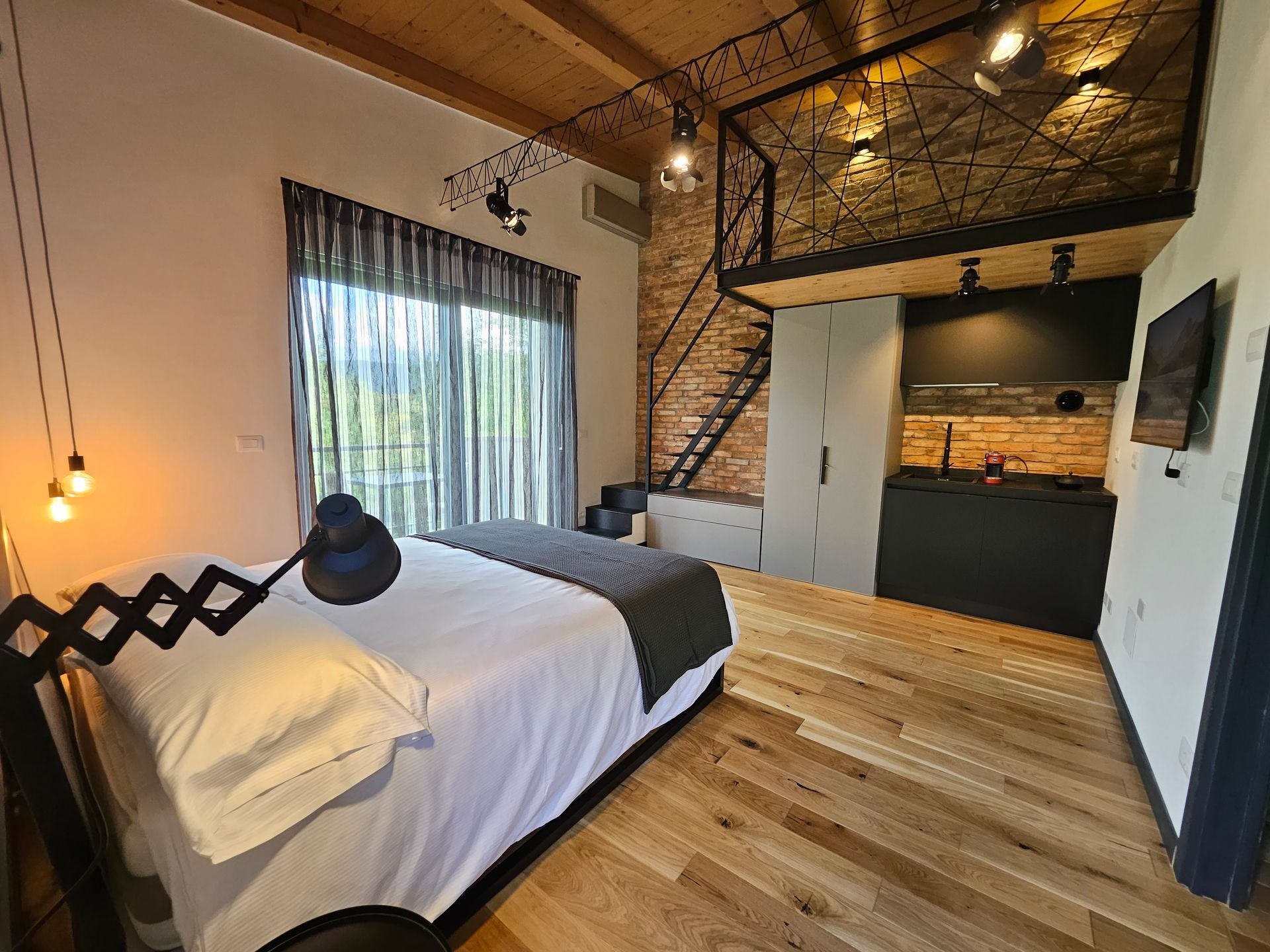 Accogliente camera da letto in stile loft con pavimento in legno, letto nero, ampia finestra e scala a chiocciola.