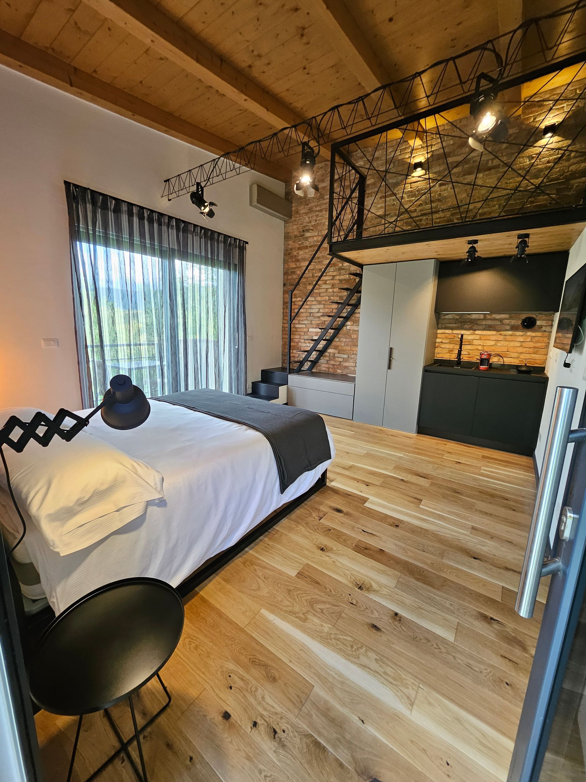 Camera da letto soppalcata con letto, sedia nera, ampia finestra, pavimento in legno e scala per il piano superiore.