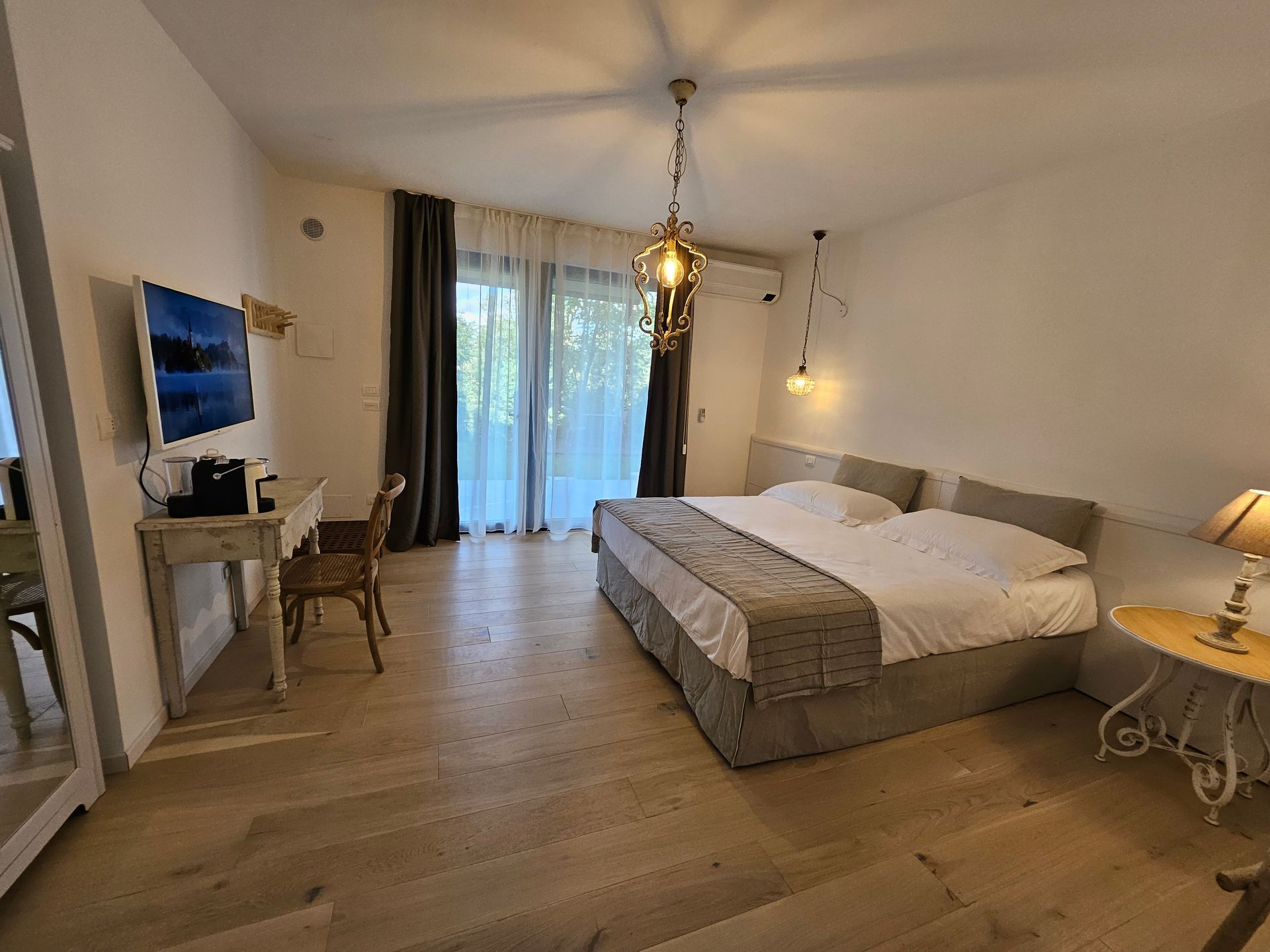 Camera da letto luminosa con un grande letto, TV su una scrivania e porte scorrevoli in vetro che conducono a un balcone.