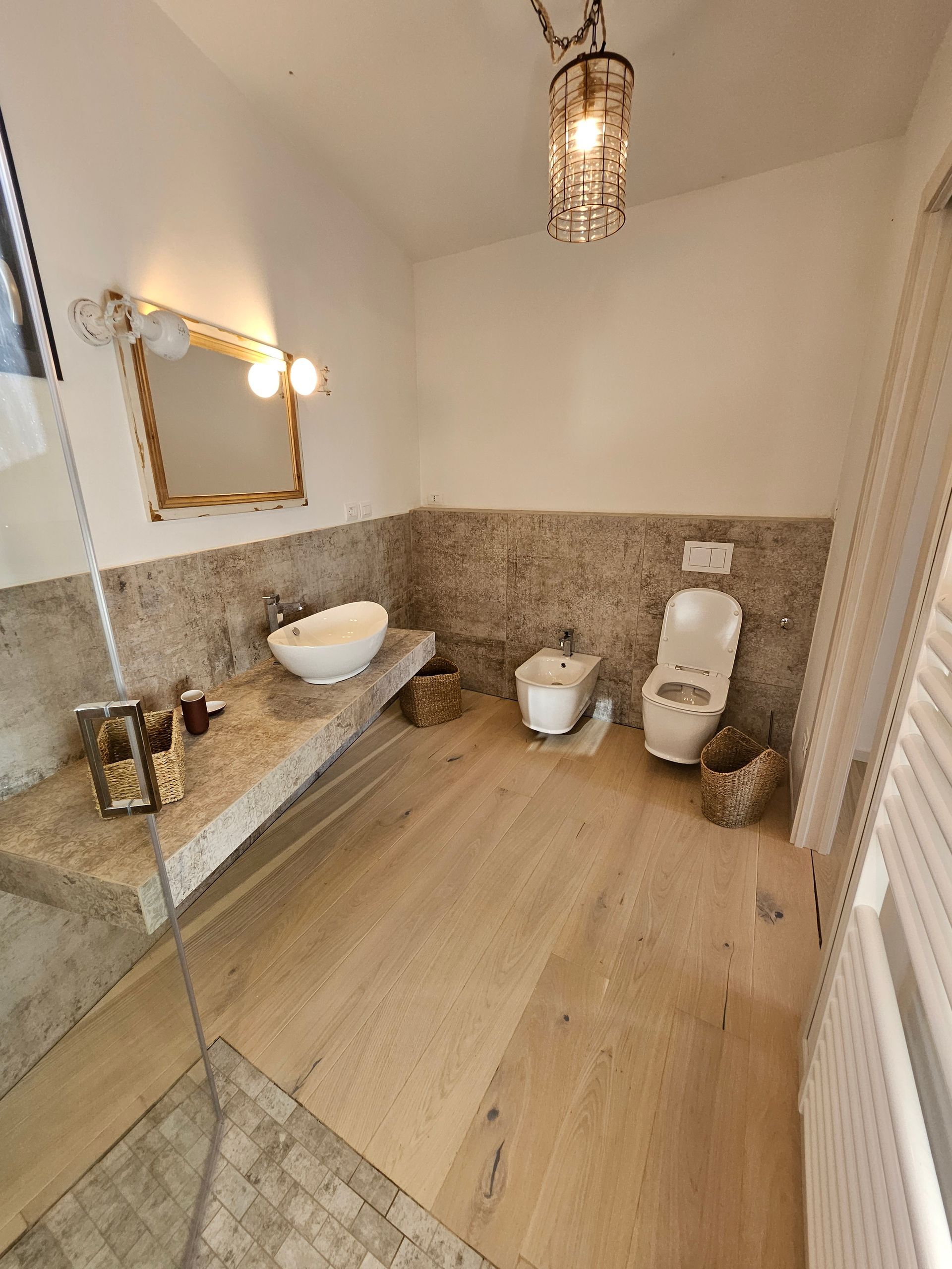 Luminoso bagno moderno con doccia in vetro, specchio a parete, lavabo sospeso, WC, bidet e pavimento effetto legno.