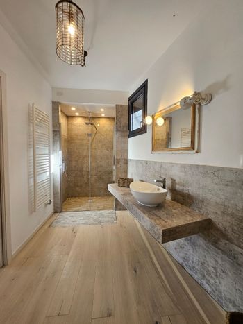Bagno moderno con doccia in pietra, mobile lavabo sospeso, specchio dorato e pavimento in legno chiaro.