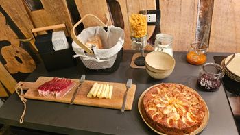 Tavolo da buffet con affettati, pane, pizza, ciotole e condimenti su un bancone nero.