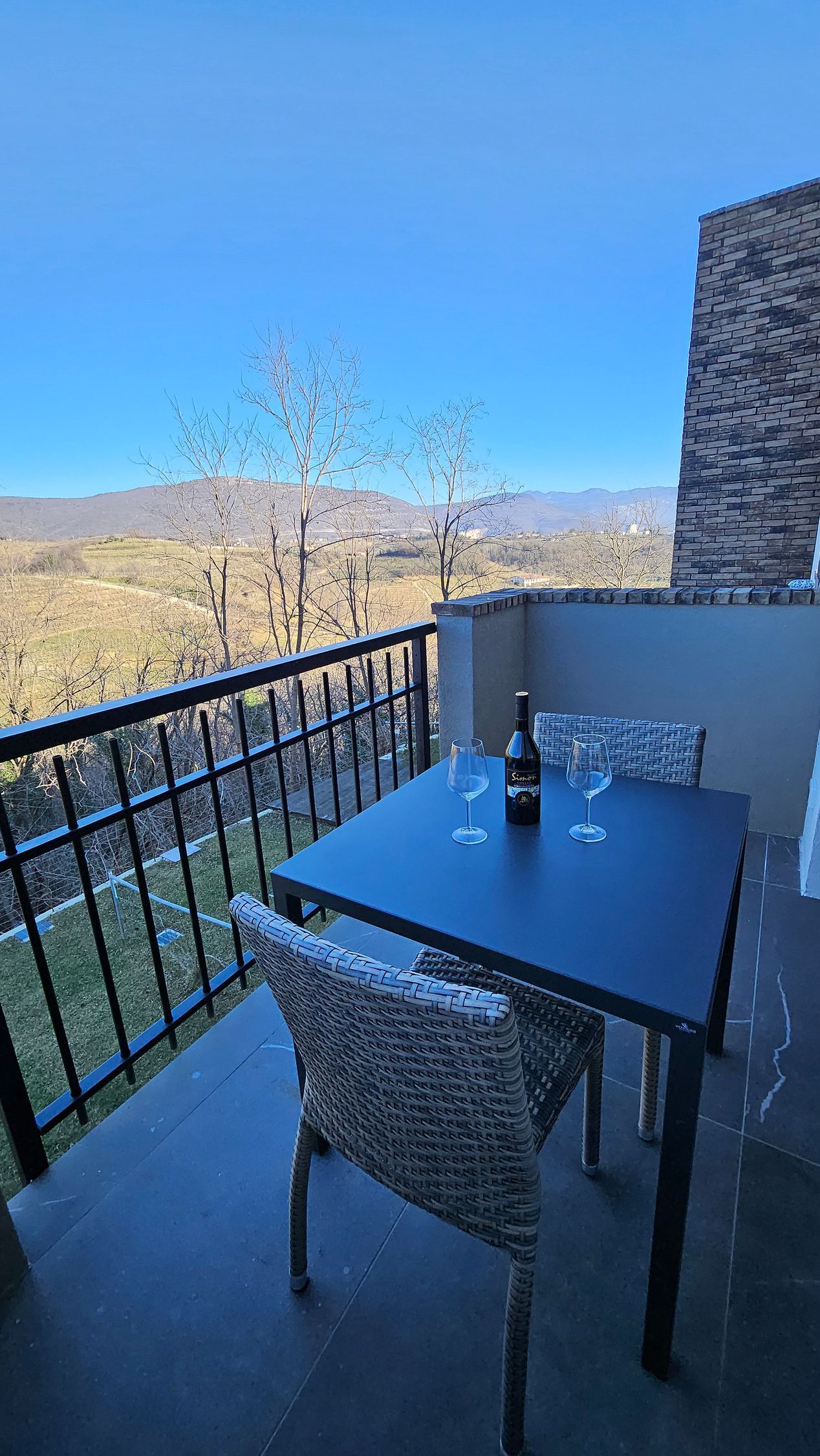 Tavolo e sedie da balcone con vista su una valle montana illuminata dal sole, sotto un cielo azzurro e limpido.