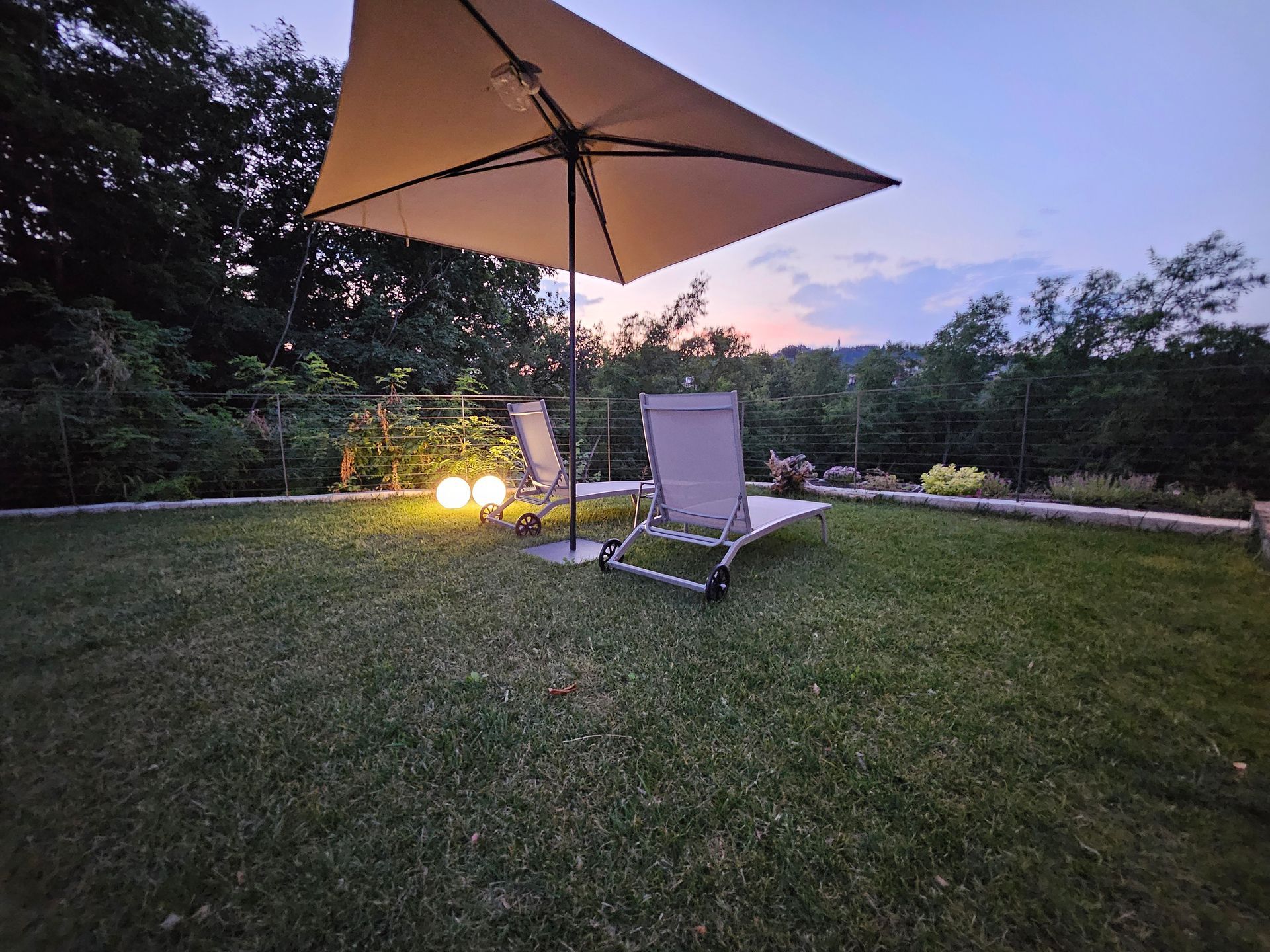 Ombrellone da patio e sdraio su un prato al crepuscolo, illuminati da calde luci da giardino.